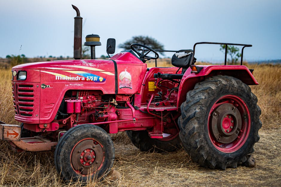 A vivid red tractor sitting idle in a dry, barren field under an overcast sky.