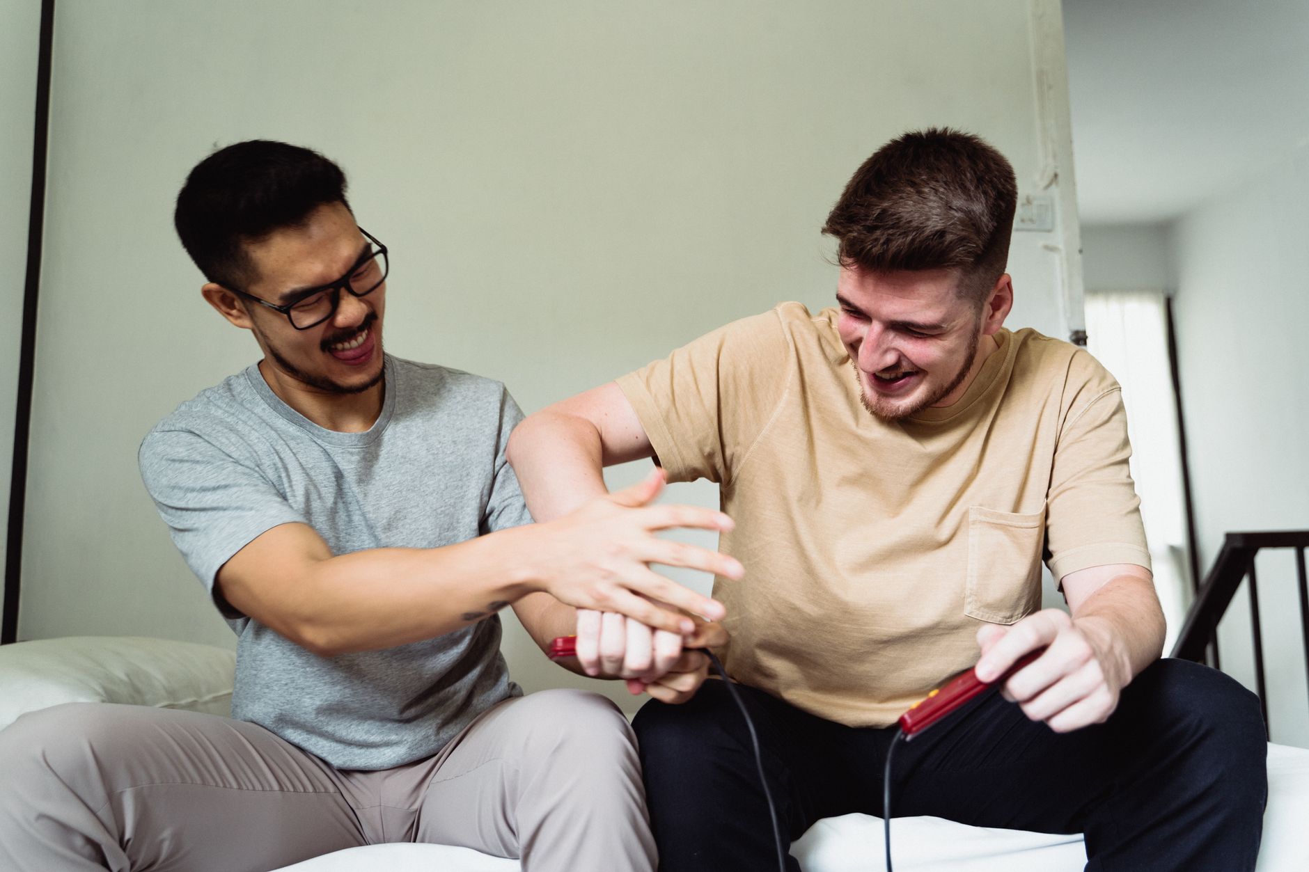 Two men laughing and playfully competing with video game controllers on a sofa. – game tester vs friend