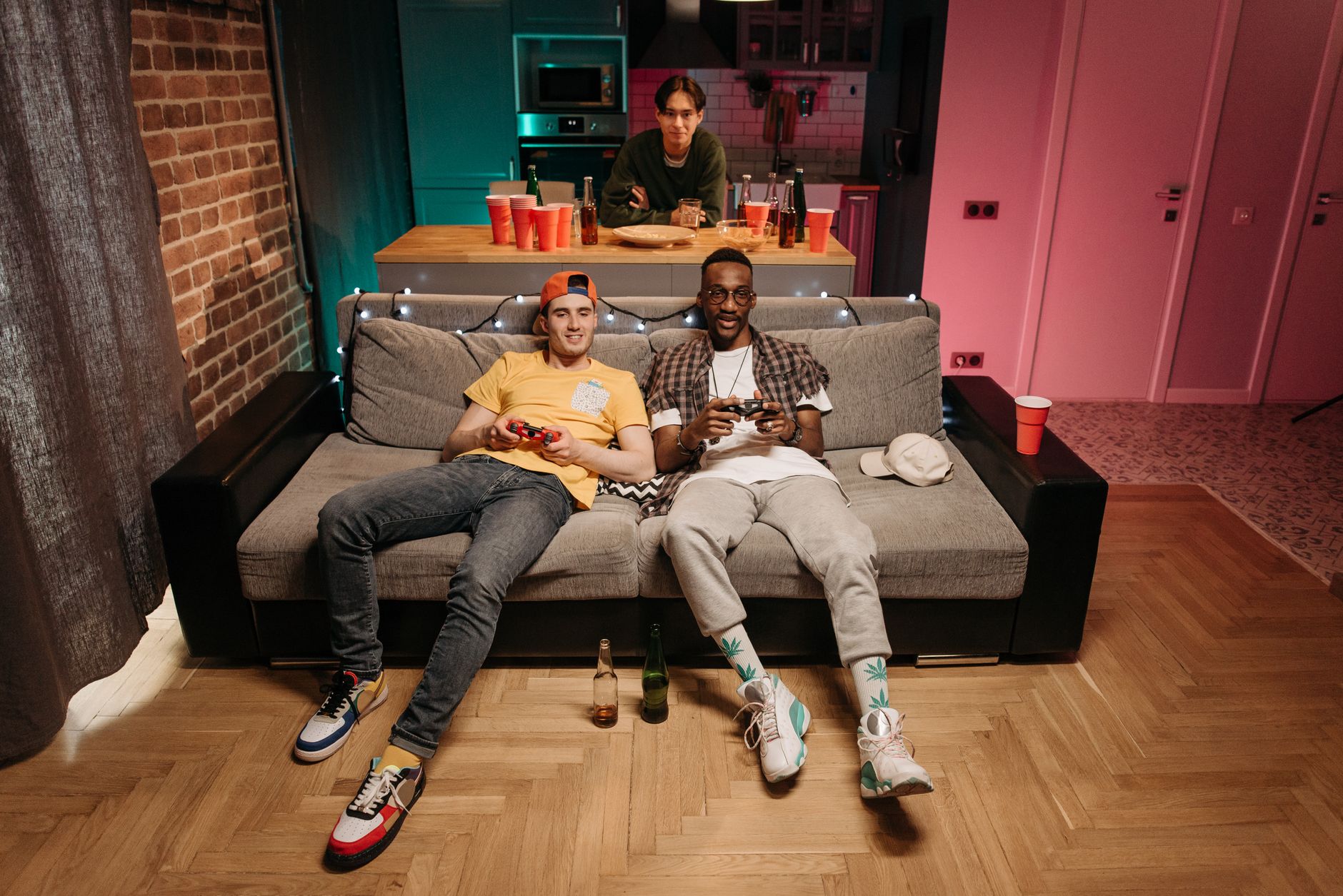 Three friends enjoying a gaming night in a cozy living room with colorful lights and drinks.