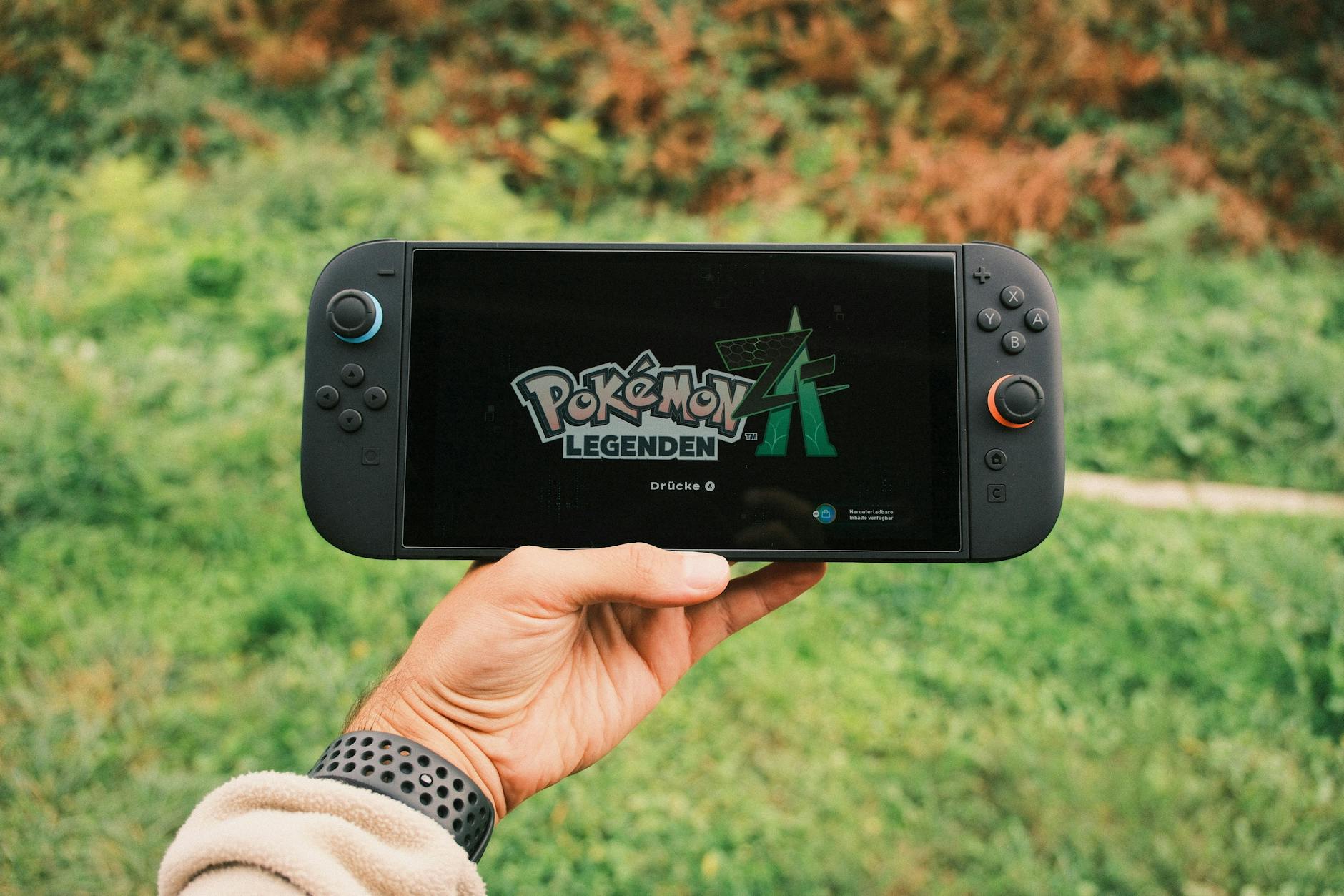 A hand holding a handheld gaming console displaying the Pokémon Legends game screen outdoors.