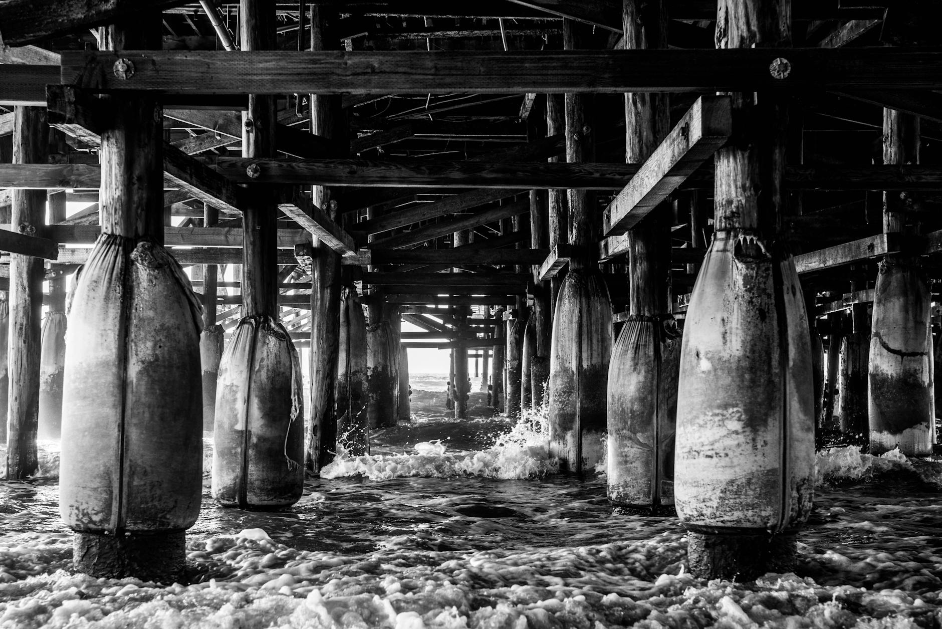 Striking black and white view showcasing waves crashing under a wooden pier with reinforcing beams.