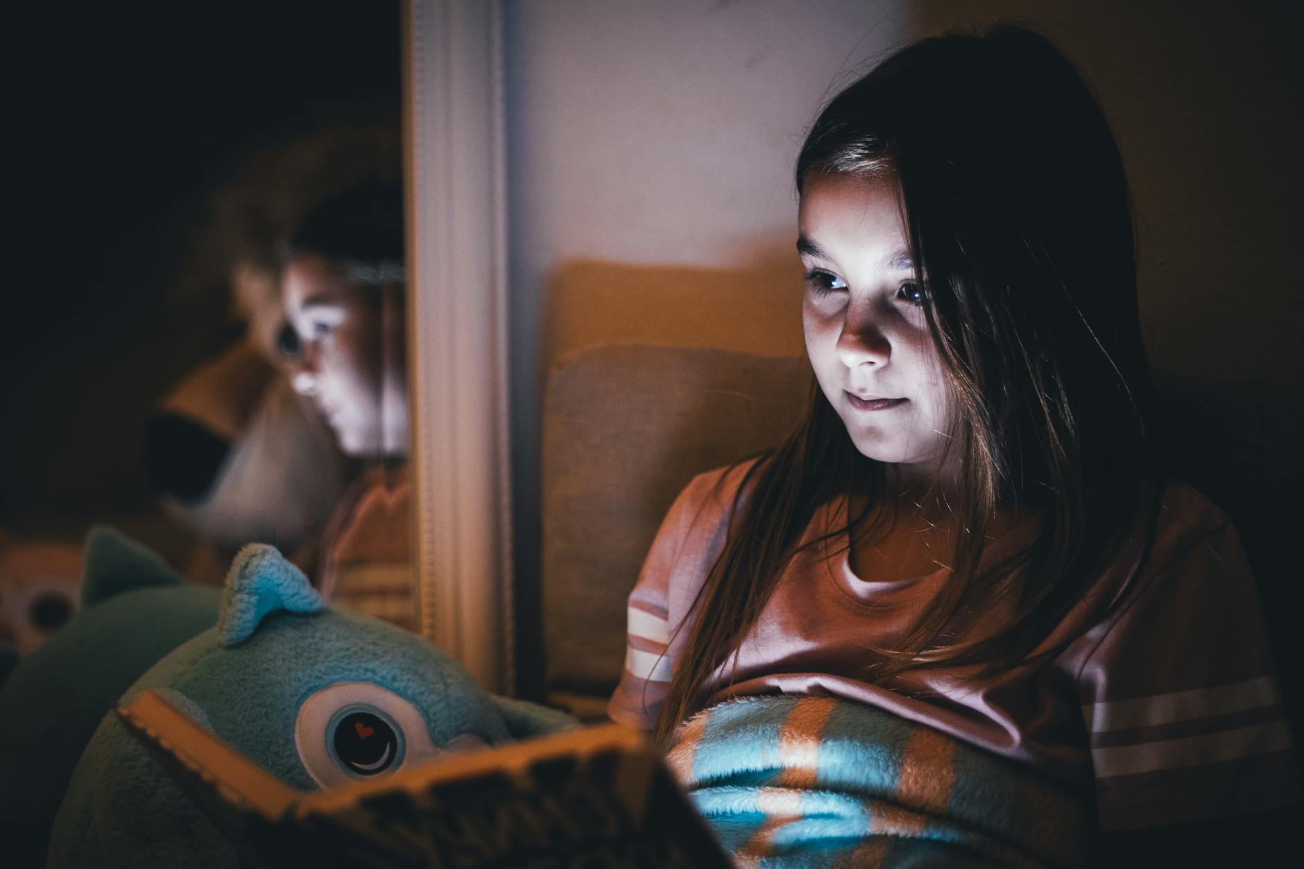 A young girl reads a book under warm light at night, reflecting on a mirror.