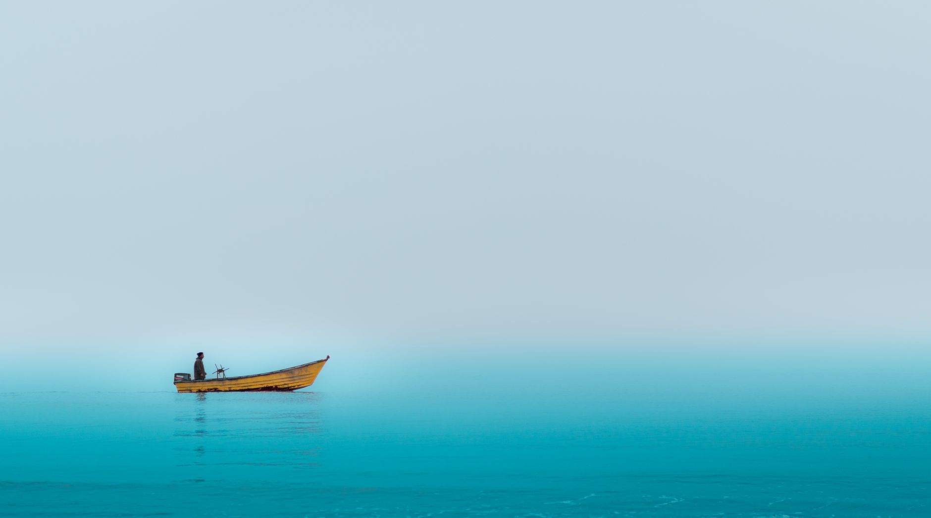 A lone fisherman stands in a boat on a misty lake, evoking tranquility.